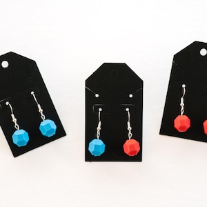 May include: Three pairs of earrings displayed on black earring cards. Each pair features a silver hook and a geometric bead. One pair has blue beads, one has red, and the third has a mix of blue and red beads. The earrings are on a white surface.