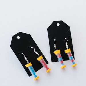 May include: Earrings featuring clear tubes filled with small red and blue beads, capped with yellow and blue plastic. The earrings hang from silver-colored fish hook ear wires, displayed on black earring cards.