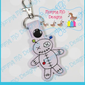 May include: A purple fabric voodoo doll keychain with a red button nose, black eyes, and stitched details. The keychain has a silver metal key ring.