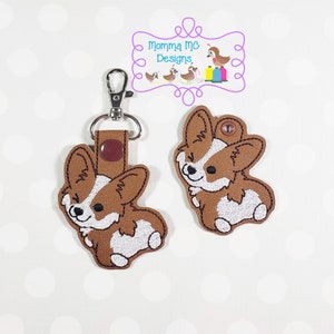 Op de afbeelding: Bruine en witte geborduurde corgi sleutelhanger met een zilveren sleutelring. De sleutelhanger is bevestigd aan een bruin leren bandje met een drukknoop. De corgi zit met zijn tong uit.