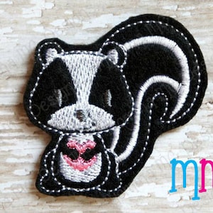May include: Black and white embroidered patch of a skunk holding a pink heart. The skunk has a black body with a white stripe down its back and a white face with black eyes.