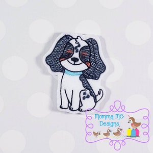 Puede incluir: Parche bordado de un perro blanco y negro con ojos marrones, sentado con un collar azul. El perro sonríe y tiene una expresión feliz.