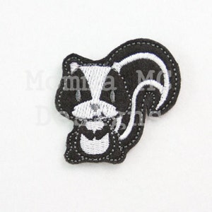 May include: A black and white skunk applique with embroidered details. The skunk has a white stripe down its back and a white face. The applique is likely for sewing or crafting projects.