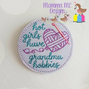 May include: A round, light purple patch with the text "hot girls have grandma hobbies" embroidered in teal and purple thread. A heart-shaped yarn ball and a pink knitting needle are also embroidered on the patch. The patch has a stitched border.