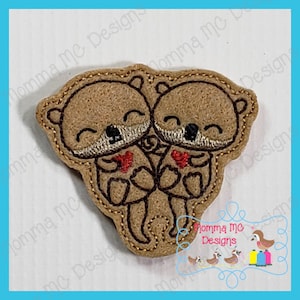 May include: Embroidered felt applique of two brown otters holding hearts. The otters are holding each other and have smiling faces.
