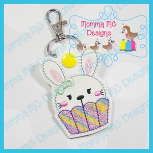 May include: A white bunny cupcake keychain with a yellow button nose, pink bow, and purple, pink, and blue striped frosting.