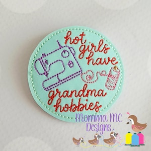 May include: A round, light teal patch with red embroidered text that reads "hot girls have grandma hobbies." A purple sewing machine and a spool of thread are also embroidered on the patch. The words "Momma mc Designs" are at the bottom.
