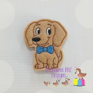 May include: A tan felt dog applique with a blue bow tie. The dog has a smiling face with black eyes and nose. The applique is likely for crafts or sewing projects. The image also includes the text "Momma mc Designs."