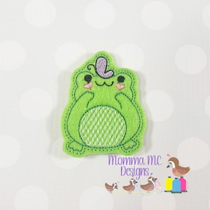 May include: Green felt applique of a frog wearing a purple flower on its head. The frog has a green and white striped body and pink cheeks.