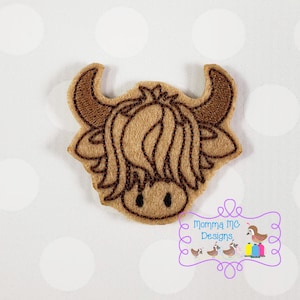 May include: A felt applique of a Highland cow's head. The cow has light brown fur, dark brown horns, and black eyes. The applique is outlined in dark brown stitching. The image includes the text "Momma MC Designs" in a purple frame.