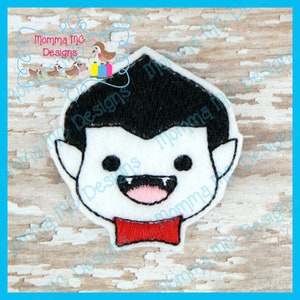 May include: A white felt vampire face with black hair, black eyes, and a red bow tie. The vampire has a smiling mouth with two fangs. The text "Momma Mc Designs" is in the upper left corner.