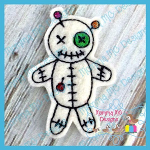 May include: White felt voodoo doll with black stitching, a red button, and a green eye. The doll has a stitched-on smile and a single pin with colorful beads on top of its head.