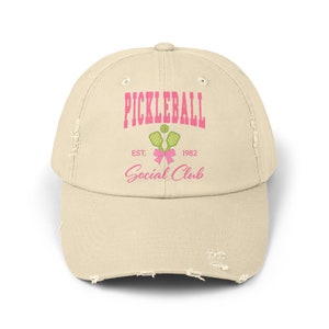 Könnte beinhalten: Eine abgenutzte beige Baseballkappe mit dem Text "Pickleball Social Club" in Pink und Grün. Der Text "EST. 1982" ist ebenfalls in Pink enthalten.