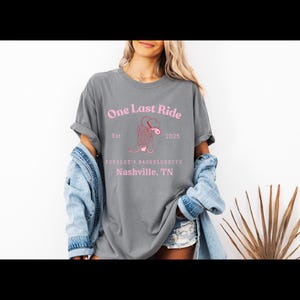 Puede incluir: Camiseta gris con texto rosa que dice "One Last Ride" con un gráfico de bota rosa y el texto "Est. 2025" y "Kensley's Bachelorette Nashville, TN".