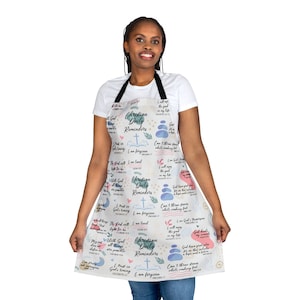 Op de afbeelding: Een witte schort met een zwarte nekband met de tekst "Christian Daily Reminders" en diverse inspirerende citaten en afbeeldingen. De schort is versierd met pastelkleuren en een kruissymbool.