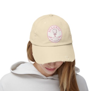 Könnte beinhalten: Eine beige, abgenutzte Baseballkappe mit einem weißen Kreislogo, das einen rosafarbenen Pickleball-Schläger und den Text "Pickleball Social Club Established 2023" zeigt.