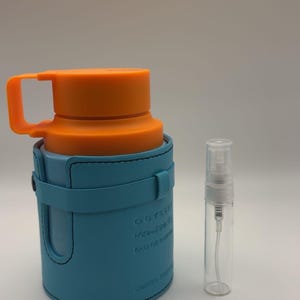 May include: A perfume bottle with an orange cap and handle, encased in a light blue holder. A small, clear spray bottle is next to it. The bottle has text that reads "ODYSSEY Mandarin Eau de Parfum Limited Edition."