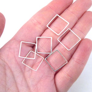 May include: Several small, open silver square charms are displayed in a hand. The charms are made of thin metal and have a simple, geometric design. They are likely used for jewelry making or other crafts.