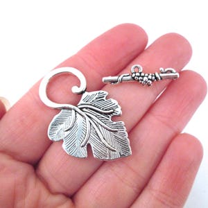 May include: Silver tone leaf and vine toggle clasp. The leaf is detailed with veins and the vine has a cluster of grapes.