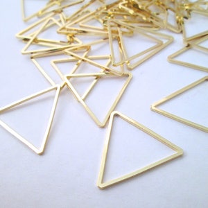 May include: A collection of small, gold-colored triangle-shaped charms. The charms are made of thin metal and have a simple, geometric design. They are scattered across a white surface, suggesting they are craft supplies or jewelry components.
