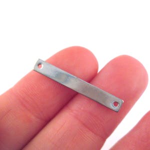 May include: A silver metal bar with two holes, held between two fingers.