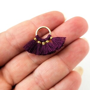 May include: A gold hoop earring with a burgundy tassel charm. The tassel is made of multiple strands of thread and has a gold bead at the top.
