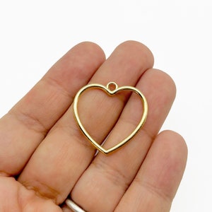 May include: Gold heart-shaped pendant with a loop at the top for hanging. The pendant is held in a hand.