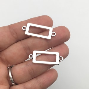 Puede incluir: Dos conectores rectangulares plateados para joyería con esquinas redondeadas y pequeños bucles en cada extremo. Los conectores se muestran sobre un fondo blanco. Los conectores miden aproximadamente 2 cm de largo.