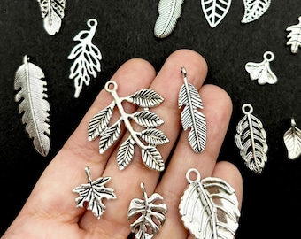 Könnte beinhalten: Eine Sammlung von silberfarbenen Metallanhängern in verschiedenen Blatt- und Federformen. Das Sortiment umfasst detaillierte botanische Designs, die von langen, schlanken Federn bis hin zu kleinen, komplexen Blättern reichen, jeweils mit einer Öse.