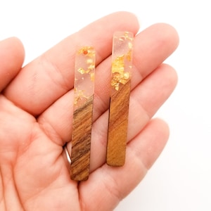 May include: Two long, rectangular earrings made of wood and clear resin with gold leaf accents. The earrings are held in a hand.