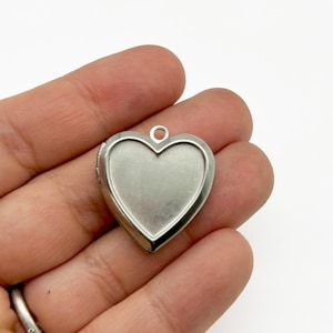 May include: Silver heart-shaped locket pendant with a hinged opening. The locket is shown in a hand.