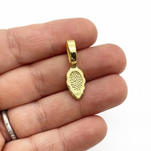 May include: A gold-colored pendant with a textured, oval-shaped design. The pendant has a small loop at the top for a chain. The pendant is held in a hand against a white background.
