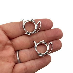 May include: Two silver-colored teapot charms. Each charm is a miniature teapot shape with a handle, spout, and lid. The charms are made of a shiny metal and are displayed against a neutral background.