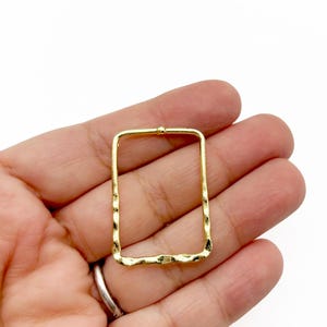 May include: A gold-colored, rectangular jewelry finding with a hammered texture. The open frame has a small loop at the top for attachment. The jewelry component is held in a hand against a white background.