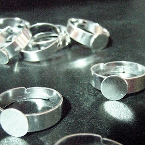 May include: A set of silver-toned adjustable ring blanks with round, flat, blank metal bases. The rings are made of metal and are designed to be used for crafting jewelry.