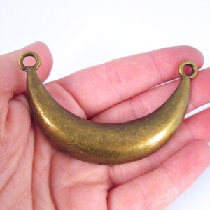 May include: A gold-toned metal crescent moon charm with two loops for hanging. The charm is smooth and has a slightly aged finish.