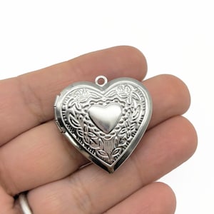 Op de afbeelding: Een zilveren hartvormige locket met een sierlijk bloemmotief. De locket heeft een kleiner hart in het midden en een lus voor een ketting. De locket wordt in een hand gehouden.