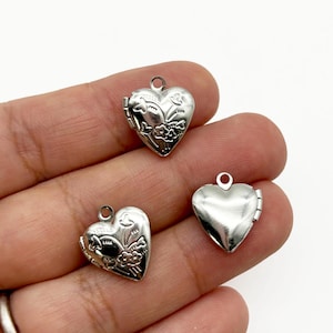May include: Three silver heart-shaped lockets. Two lockets feature floral designs, while the third is plain. Each locket has a small loop at the top for attaching to a chain or bracelet. The lockets are small and delicate.