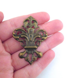 May include: A gold-toned metal fleur-de-lis design with intricate details. The design is ornate and has a vintage look.