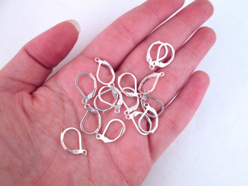 May include: A close-up of a hand holding a pile of silver earring hooks. The hooks are made of metal and have a teardrop shape.