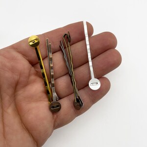May include: Four hair clips with different finishes: gold, bronze, silver, and a wavy bronze clip. Each clip has a round button on the end.