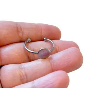May include: A silver-colored adjustable ring with a small, round, light pink stone. The ring has a simple, minimalist design with a curved band and small ball accents on each end. The stone is set in the center of the ring.