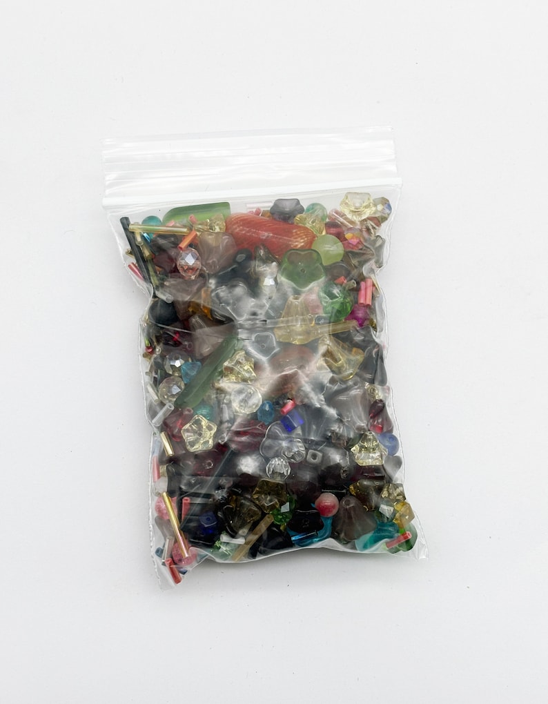 May include: A clear plastic bag filled with a variety of colorful beads. The beads are made of glass and plastic and come in a variety of shapes and sizes. The beads are a mix of red, green, blue, yellow, and clear.