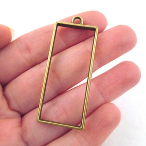 May include: An antique brass rectangular pendant frame. The open frame has a small loop at the top for attaching to a chain or cord. The pendant is held in a hand.
