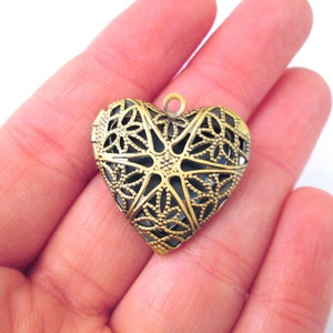 Puede incluir: Un colgante de metal dorado en forma de corazón con un intrincado diseño de filigrana. El colgante tiene un bucle en la parte superior para sujetar una cadena.