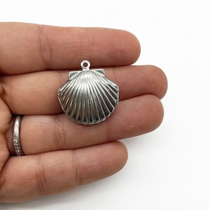 May include: Silver seashell charm pendant with a detailed, ribbed texture. The pendant is openable and has a loop at the top for attaching to a chain or bracelet.