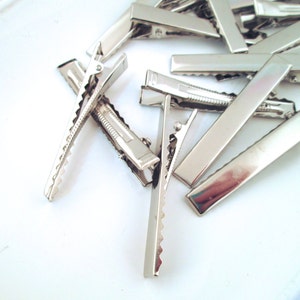 May include: A group of silver alligator clips, also known as hair clips, are scattered on a white surface. The clips are all the same size and shape, and they have a smooth, shiny finish.
