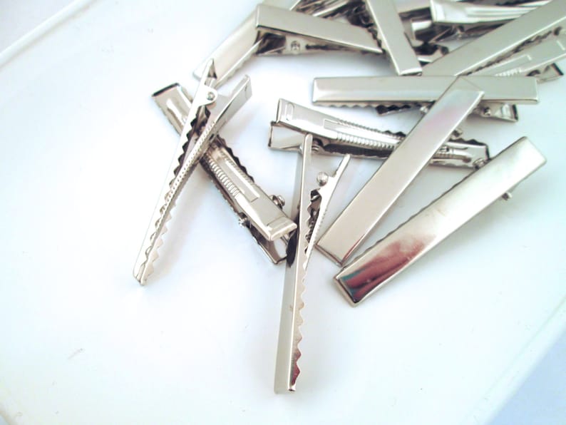 May include: A group of silver alligator clips, also known as hair clips, are scattered on a white surface. The clips are all the same size and shape, and they have a smooth, shiny finish.