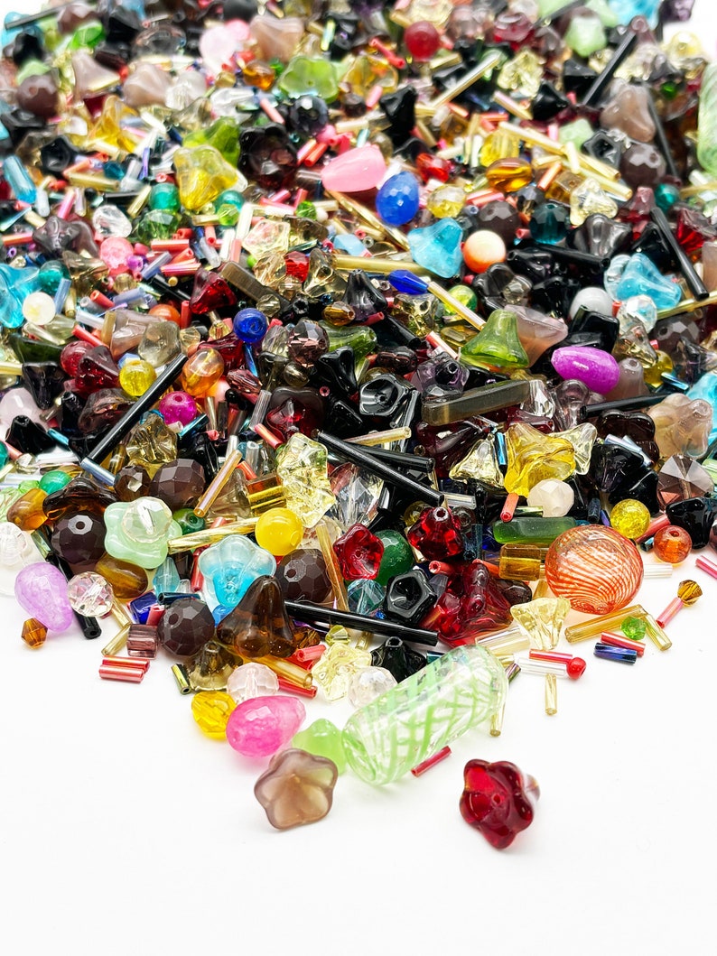 May include: A large assortment of colorful glass beads in various shapes and sizes, including round, star, and teardrop shapes. The beads are scattered on a white surface.