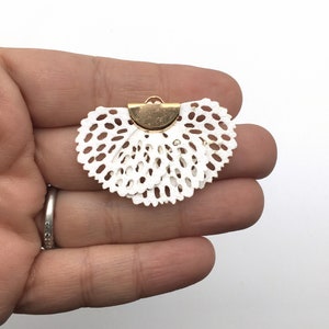 May include: White lace trim with a gold metal clasp. The lace is in a fan shape.